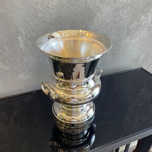 Puede incluir: Un recipiente plateado ornamentado, con un borde ancho y acampanado y una base bulbosa. El recipiente tiene dos asas peque&ntilde;as y una superficie reflectante, lo que sugiere que est&aacute; hecho de metal pulido. El dise&ntilde;o es cl&aacute;sico y elegante.