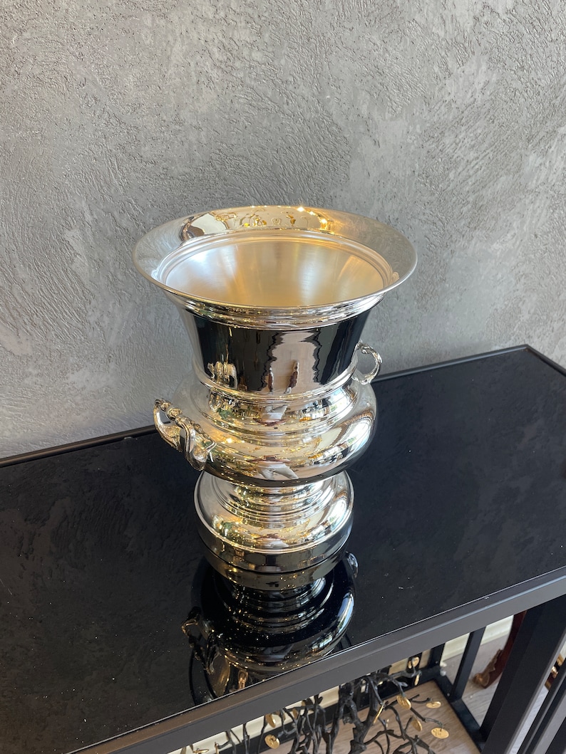 Puede incluir: Un recipiente plateado ornamentado, con un borde ancho y acampanado y una base bulbosa. El recipiente tiene dos asas peque&ntilde;as y una superficie reflectante, lo que sugiere que est&aacute; hecho de metal pulido. El dise&ntilde;o es cl&aacute;sico y elegante.