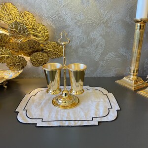 May include: Gold metal stand with two gold metal cups. The stand has a decorative design and is on a white linen placemat with a black border.