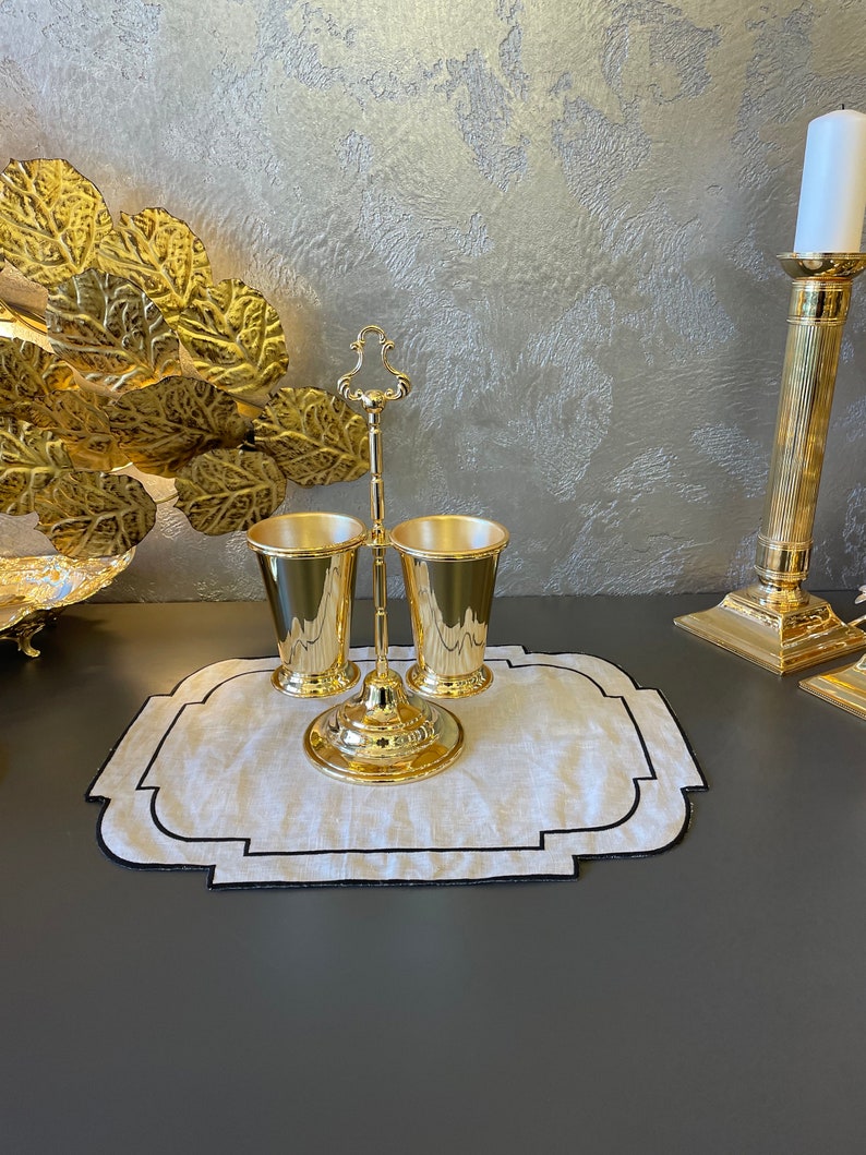 May include: Gold metal stand with two gold metal cups. The stand has a decorative design and is on a white linen placemat with a black border.