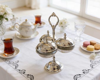 May include: A silver-toned metal tiered serving stand featuring two round bowls with matching domed lids and a central decorative handle. The stand has a circular base and is positioned on a white tablecloth with black embroidered floral borders.