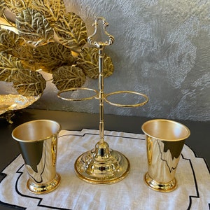May include: A gold metal stand with two rings for holding glasses. Two gold metal cups are on a white linen placemat with a black border.