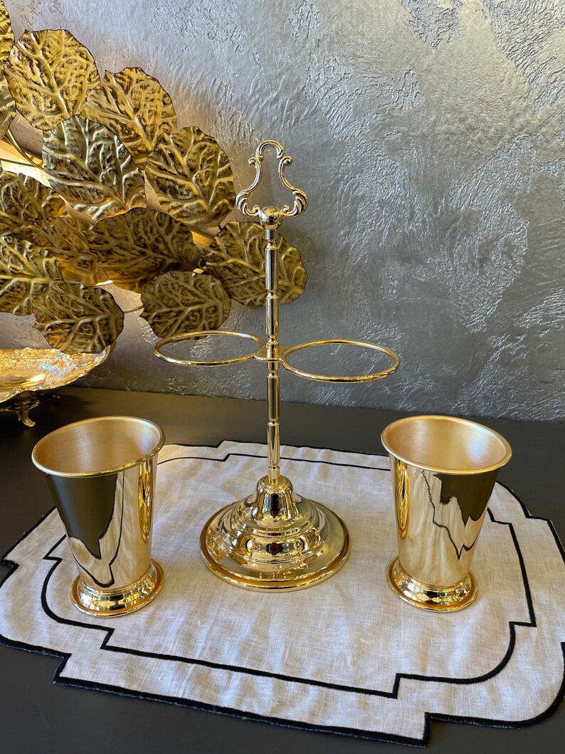 May include: A gold metal stand with two rings for holding glasses. Two gold metal cups are on a white linen placemat with a black border.