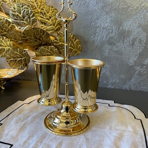 May include: Gold metal stand with three cups. The stand has a decorative handle and a round base. The cups are smooth and shiny.