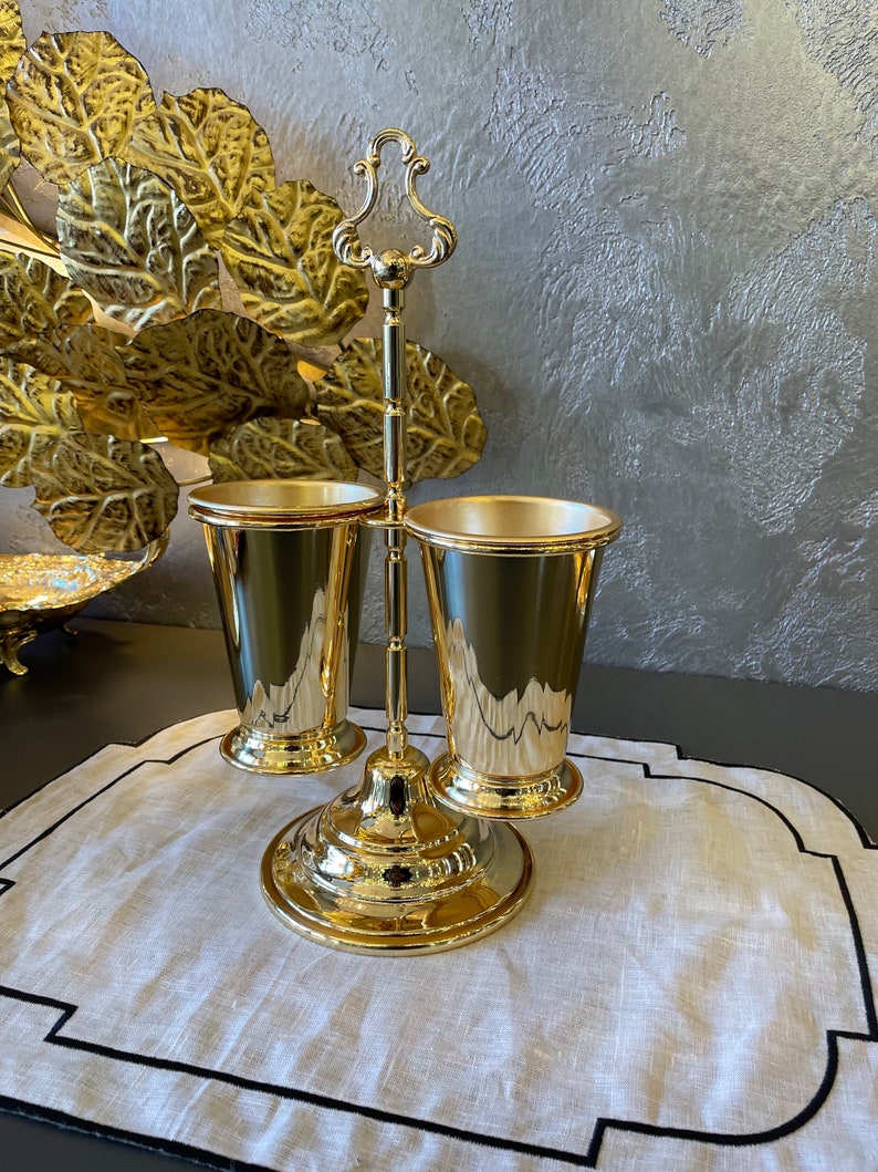 May include: Gold metal stand with three cups. The stand has a decorative handle and a round base. The cups are smooth and shiny.
