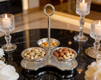 May include: A silver-toned metal serving stand featuring three circular bowls arranged in a clover-like pattern. The stand includes a central vertical handle with a ring at the top. The bowls contain various nuts, including pistachios and almonds.