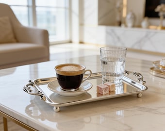 May include: A rectangular silver-toned serving tray with ornate handles and small ball feet. On the tray sits a ceramic cup of dark coffee on a matching saucer, a clear glass of water, and two small, light pink square-shaped confectioneries.