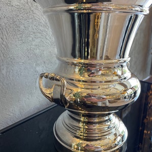 Puede incluir: Un jarr&oacute;n de metal plateado pulido con una parte superior ancha y acampanada y una base bulbosa. El jarr&oacute;n tiene dos asas peque&ntilde;as y refleja el entorno. El jarr&oacute;n probablemente se utiliza con fines decorativos.