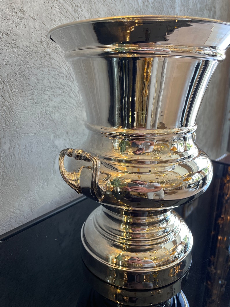 Puede incluir: Un jarr&oacute;n de metal plateado pulido con una parte superior ancha y acampanada y una base bulbosa. El jarr&oacute;n tiene dos asas peque&ntilde;as y refleja el entorno. El jarr&oacute;n probablemente se utiliza con fines decorativos.