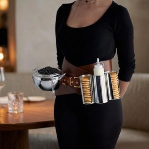 May include: A brown leather utility belt worn around the waist, featuring a silver metal bowl filled with black caviar, two silver cylindrical holders containing stacks of small round pancakes, and a white squeeze bottle of cream.
