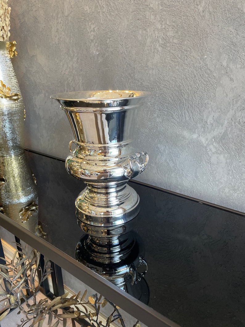 Puede incluir: Una urna decorativa plateada pulida con un borde ancho y acampanado y asas ornamentadas. La urna tiene una secci&oacute;n media bulbosa y una base robusta. La superficie reflectante de la urna crea un efecto de espejo. La urna se exhibe sobre una superficie negra.