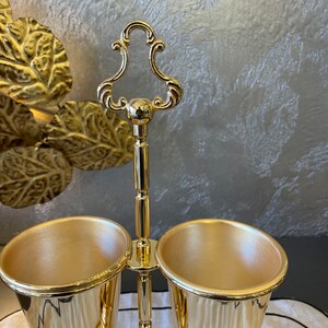 May include: A gold metal utensil holder with two gold metal cups. The holder has a decorative top with a loop handle.