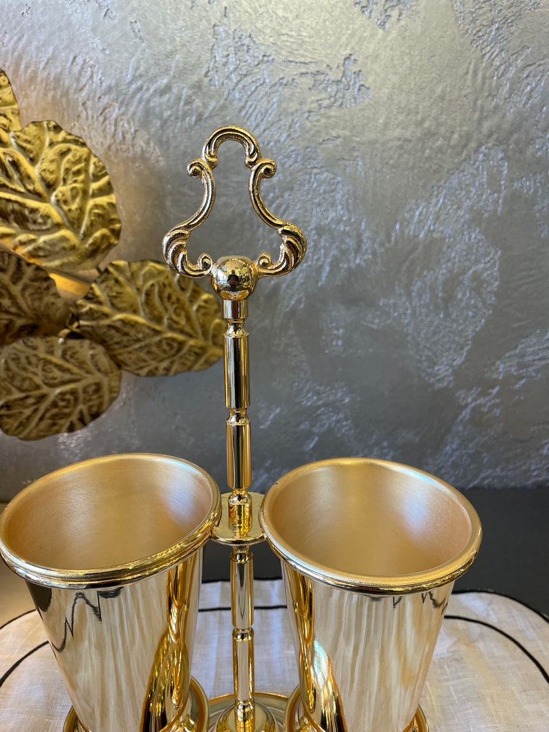 May include: A gold metal utensil holder with two gold metal cups. The holder has a decorative top with a loop handle.