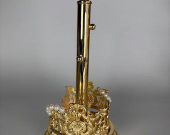 May include: A gold-colored metal jewelry display stand featuring a central vertical rod with two small pegs, set upon a circular base. The base is surrounded by an ornate, crown-shaped decorative border embellished with small white faux pearls.