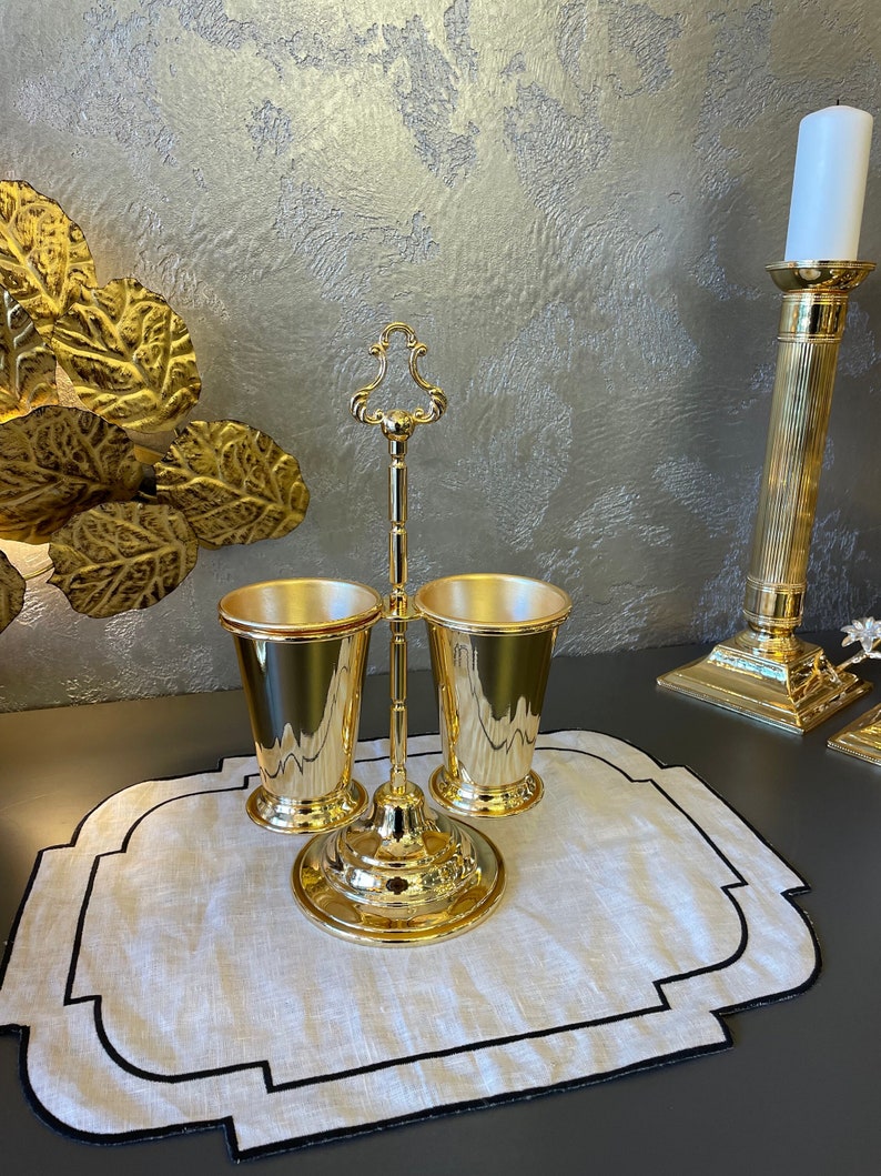 May include: Gold metal stand with two gold cups. The stand has a decorative top and a round base. The cups are on a white linen placemat with a black border.