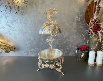 May include: A gold-toned metal serving dish featuring a bowl base supported by three legs adorned with floral accents. A matching domed lid is suspended above the bowl by a central stem, which is also decorated with delicate metal flowers and vines.