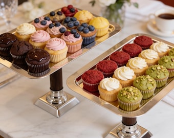 May include: Two tiered silver serving stands with dark wood pedestals holding an assortment of cupcakes. The left stand features chocolate, pink frosted, and blueberry topped cupcakes. The right stand displays red velvet, white frosted, and pistachio cupcakes.