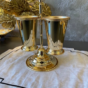 May include: Gold metal stand with two gold metal cups. The stand has a round base and a vertical rod with two arms that hold the cups.