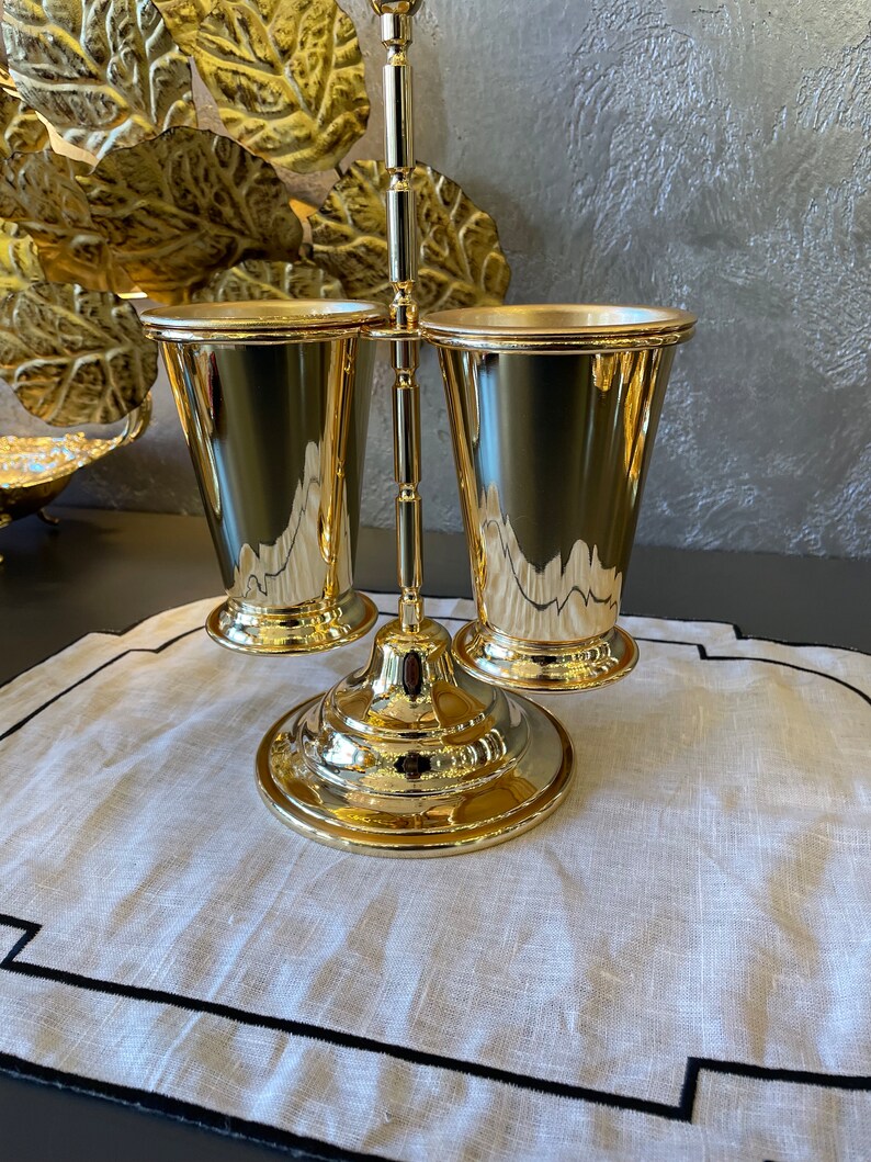 May include: Gold metal stand with two gold metal cups. The stand has a round base and a vertical rod with two arms that hold the cups.