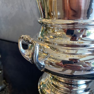 Puede incluir: Un cubo de champ&aacute;n de metal plateado brillante con un asa decorativa. El cubo tiene una parte superior ancha y acanalada y una base bulbosa. El asa tiene un dise&ntilde;o detallado. La superficie refleja el entorno.