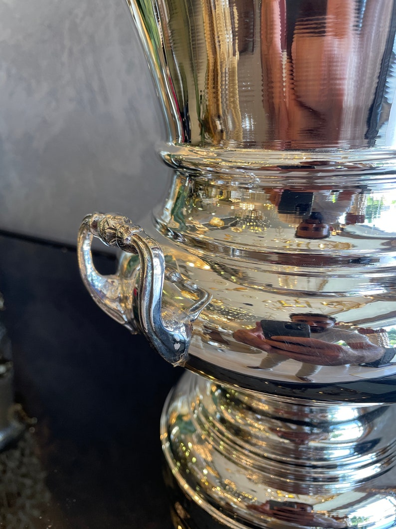Puede incluir: Un cubo de champ&aacute;n de metal plateado brillante con un asa decorativa. El cubo tiene una parte superior ancha y acanalada y una base bulbosa. El asa tiene un dise&ntilde;o detallado. La superficie refleja el entorno.