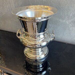 Puede incluir: Un jarr&oacute;n decorativo de metal plateado pulido con un borde ancho y acampanado y una base bulbosa. El jarr&oacute;n presenta dos asas peque&ntilde;as y una superficie reflectante, creando una est&eacute;tica moderna y elegante. El jarr&oacute;n mide unos 30 cm de altura.