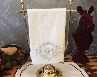 以下が含まれることがあります： 刺繍のデザインと小さなピンクのリボンが付いた白いタオルが付いた、金色のタオル掛け。 金色のベルが、黒い縁取りのある白いプレースマットの上に置かれています。 背景には、バーガンディのベルベットのロバのバストがあります。