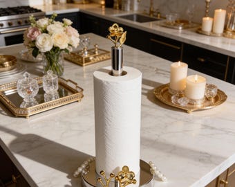 May include: A silver paper towel holder featuring a cylindrical base and a vertical rod topped with a gold-colored metal flower finial. The base is decorated with a gold-colored metal floral branch accent and a string of white pearls along the edge.