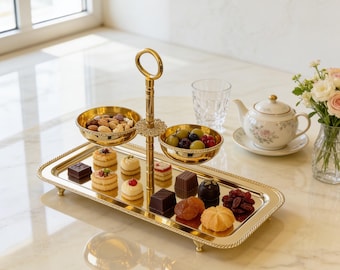 May include: A gold-colored, two-tiered serving tray with a handle, holding nuts, grapes, and various pastries. A teapot, glass, and vase of flowers are also on the marble surface, creating an elegant presentation.