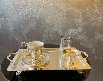 May include: A rectangular silver-toned serving tray with ornate handles and feet, holding a white porcelain cup with a silver filigree holder, a matching saucer, a clear glass with a silver rim, and a small silver pedestal dish.