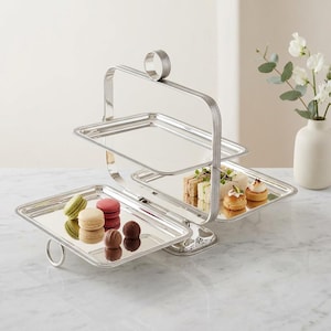 May include: A three-tiered silver serving tray with a handle. The tray is filled with macarons, sandwiches, and small pastries. The handle has a ring at the top for easy carrying. The tray is on a marble surface.