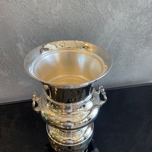 Puede incluir: Una cubitera plateada con un borde ancho y acanalado y dos asas ornamentadas. La cubitera tiene una superficie pulida y reflectante y una base escalonada. El interior es liso y vac&iacute;o.
