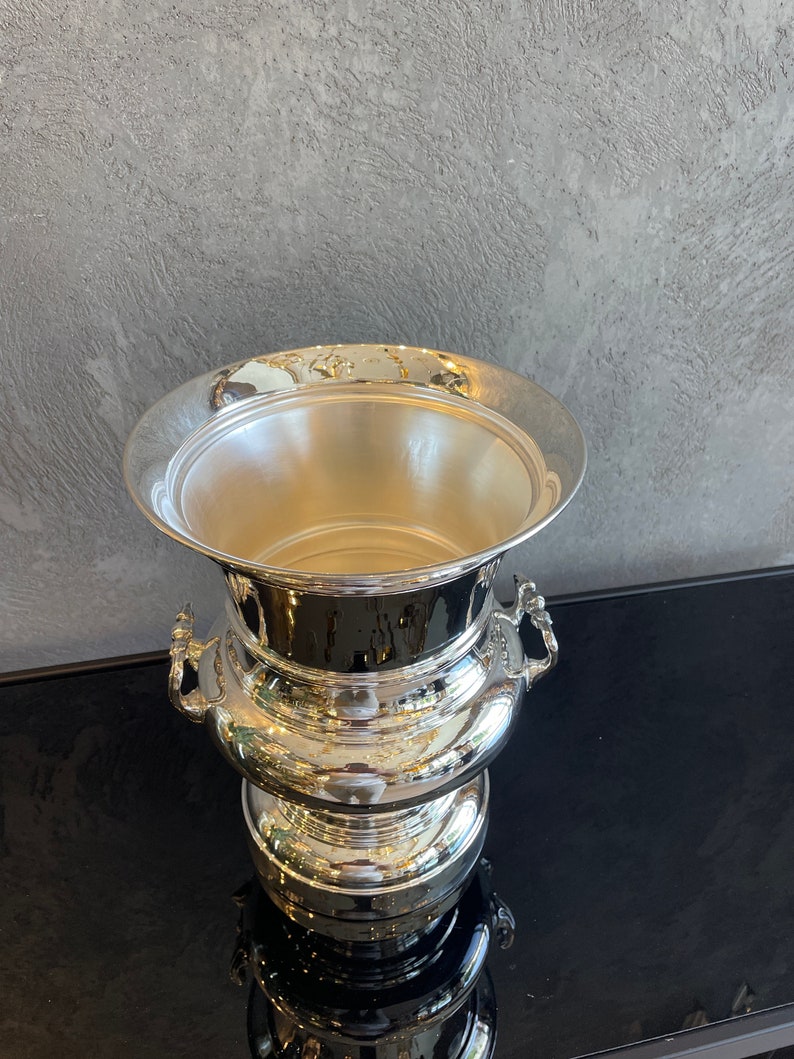 Puede incluir: Una cubitera plateada con un borde ancho y acanalado y dos asas ornamentadas. La cubitera tiene una superficie pulida y reflectante y una base escalonada. El interior es liso y vac&iacute;o.