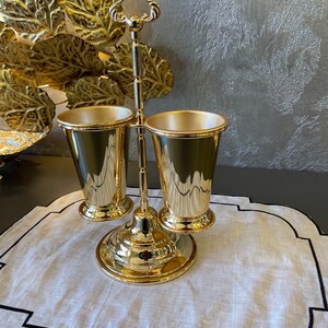 May include: Gold metal stand with two gold metal cups. The stand has a decorative handle on top and a round base. The cups are placed on the stand and are facing each other. The stand is on a white linen placemat with a black border.
