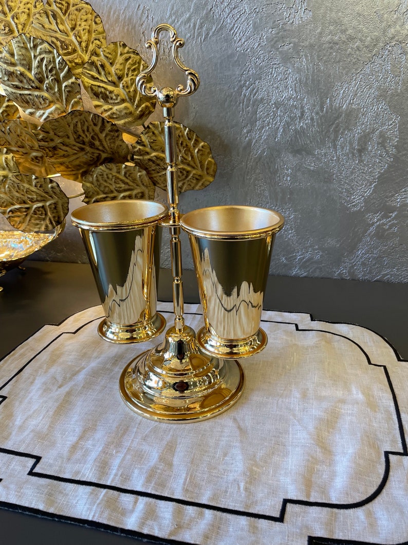 May include: Gold metal stand with two gold metal cups. The stand has a decorative handle on top and a round base. The cups are placed on the stand and are facing each other. The stand is on a white linen placemat with a black border.