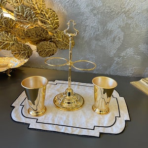May include: A gold metal stand with two rings for holding glasses, and two gold metal cups on a white linen placemat with a black border.