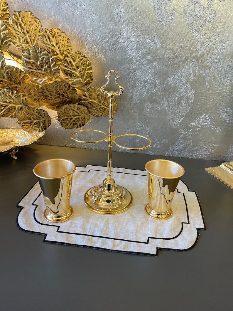 May include: A gold metal stand with two rings for holding glasses, and two gold metal cups on a white linen placemat with a black border.