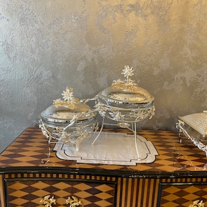 Könnte beinhalten: Silberne Servierschalen mit floralen Details und goldenen Akzenten. Die Schalen stehen auf einem dekorativen Ständer mit einer schwarz-weiß gemusterten Matte. Der Schrank hat ein braunes und goldenes Rautenmuster.