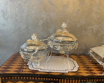 Op de afbeelding: Zilveren serveerschalen met bloemendetails en gouden accenten. De schalen staan op een decoratieve standaard met een zwart-wit patroonmat. De kast heeft een bruin en goud diamantpatroon.