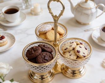 May include: A two-tiered serving stand featuring two clear, diamond-patterned glass bowls with gold-toned metal rims and a central gold-toned handle. The bowls are filled with assorted round cookies, including chocolate chip and cranberry varieties.