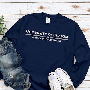 Könnte beinhalten: Ein marineblauer Pullover mit dem Text "University of Custom Est. 1852 School of Engineering" in Weiß.