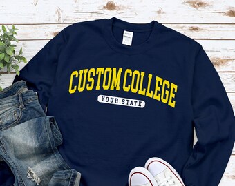 Pode incluir: Camisola azul marinho com texto amarelo que diz "CUSTOM COLLEGE" e "YOUR STATE".