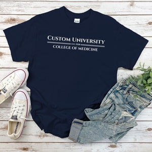 May include: A navy blue t-shirt with white text that reads "Custom University Est. 1958 College of Medicine". The shirt is laid flat on a white wooden surface with a pair of white sneakers and blue jeans.
