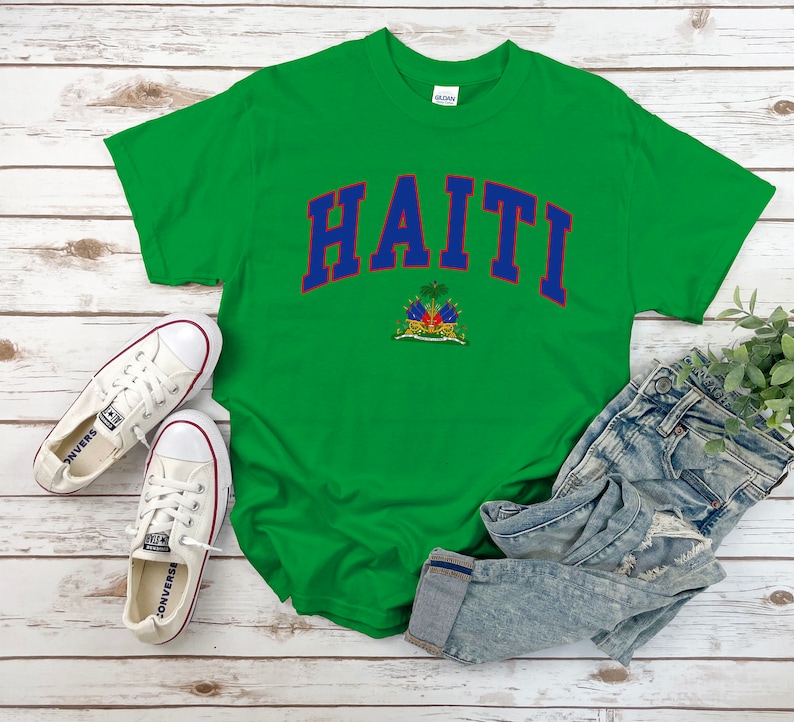 Op de afbeelding: Groen T-shirt met het woord "HAITI" in blauwe en rode letters met het embleem van de Ha&iuml;tiaanse vlag eronder.