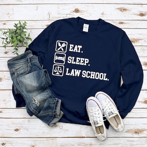 Può includere: Felpa blu navy con la scritta bianca "EAT. SLEEP. LAW SCHOOL." e icone. L'immagine mostra anche jeans blu e scarpe da ginnastica bianche. Un capo d'abbigliamento casual.
