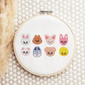 Può includere: Un telaio da ricamo a punto croce con otto facce di animali dei cartoni animati in vari colori. Gli animali includono un coniglio, un orso, una volpe e un pulcino. Il ricamo è ambientato su uno sfondo di tessuto bianco, incorniciato da un telaio di legno.