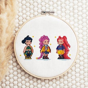 May include: A cross-stitch embroidery hoop featuring three pixelated figures with dark hair, pink hair, and red hair. The figures are wearing various outfits, including a green top, a yellow jacket, and a blue top. The background is white.