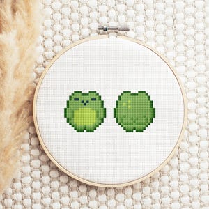 Puede incluir: Un bastidor de bordado de punto de cruz con dos ranas verdes pixeladas. Las ranas están en una pose relajada, una mirando hacia adelante y la otra hacia un lado. El bordado está sobre un fondo de tela blanca.