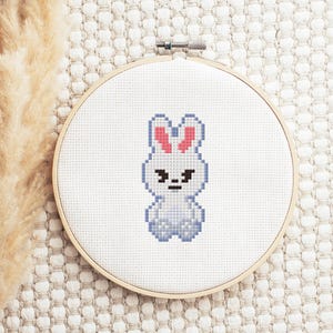 May include: A cross-stitch embroidery hoop featuring a pixelated bunny design. The bunny is white with pink ears and a stern expression. The embroidery is set against a white fabric background, framed by a wooden hoop.