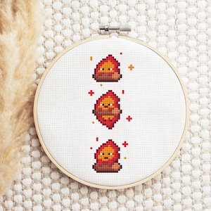 May include: A cross-stitch embroidery hoop featuring three pixelated fire designs in shades of red, orange, and brown. The embroidery is set against a white fabric background, with a wooden hoop. Decorative accents include small red and yellow crosses.